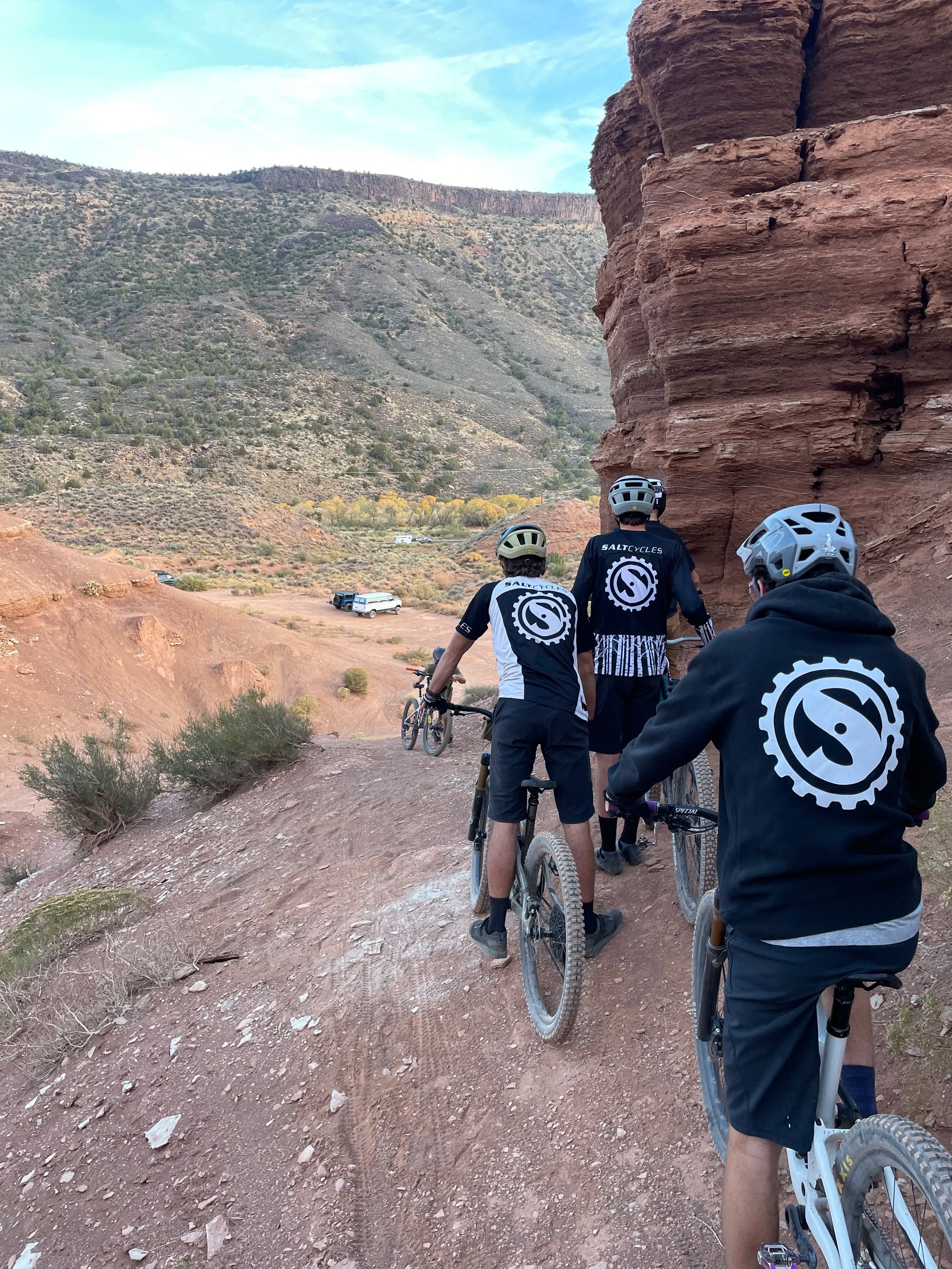 Three mountain bikers on a trail with rocky cliffs and desert landscape in the background.